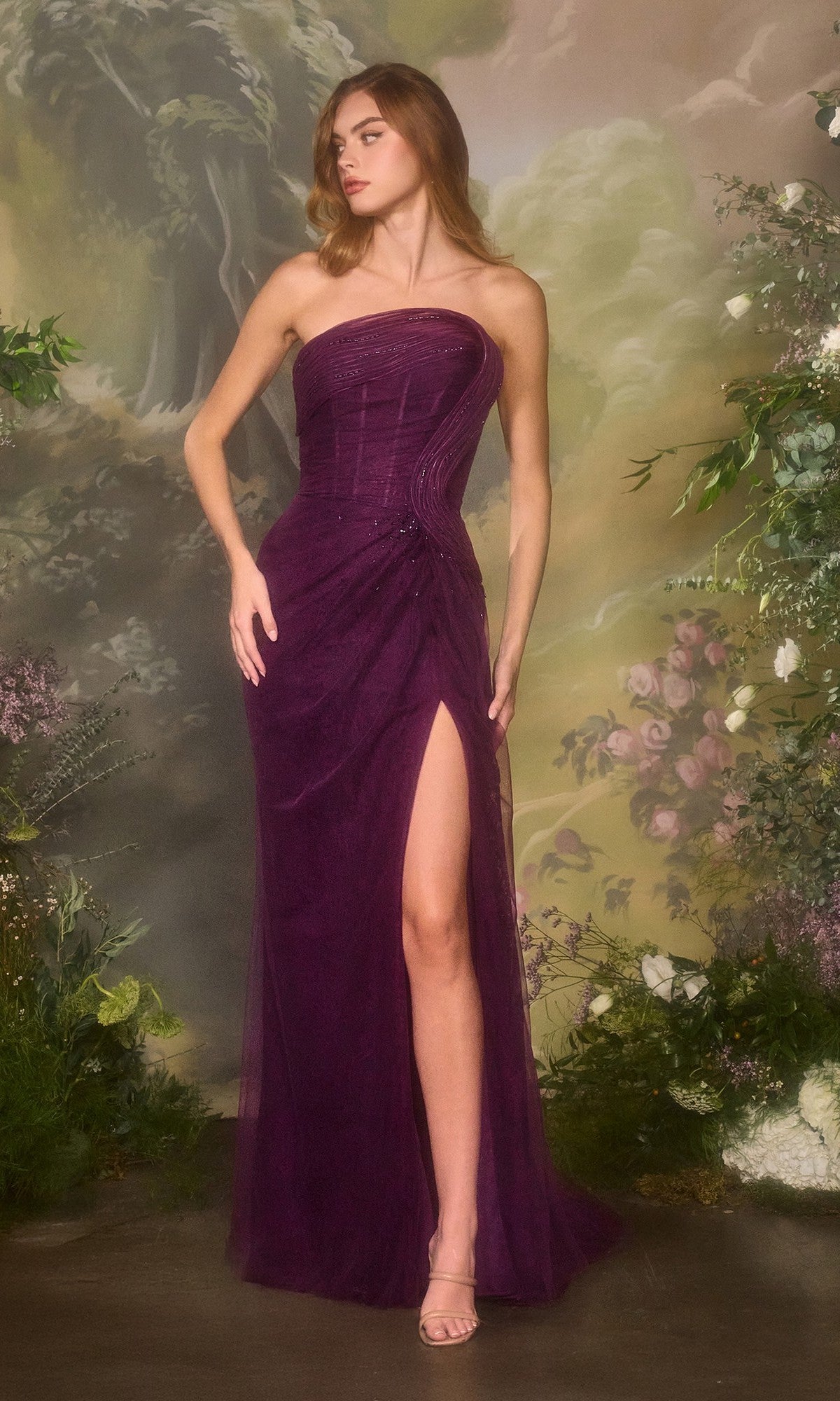 A woman stands before a painted floral backdrop in the Andrea and Leo A1497 long prom dress, a strapless, floor-length eggplant gown with a thigh-high slit.
